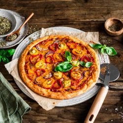 Eine hausgemachte, proteinreiche Pizza mit gelbem und rotem Tomatenbelag, Basilikumblättern und Kräutern steht auf einem rustikalen Holztisch. Daneben stehen ein Pizzaschneider, eine Serviette und kleine Schüsseln mit Gewürzen und Salz.