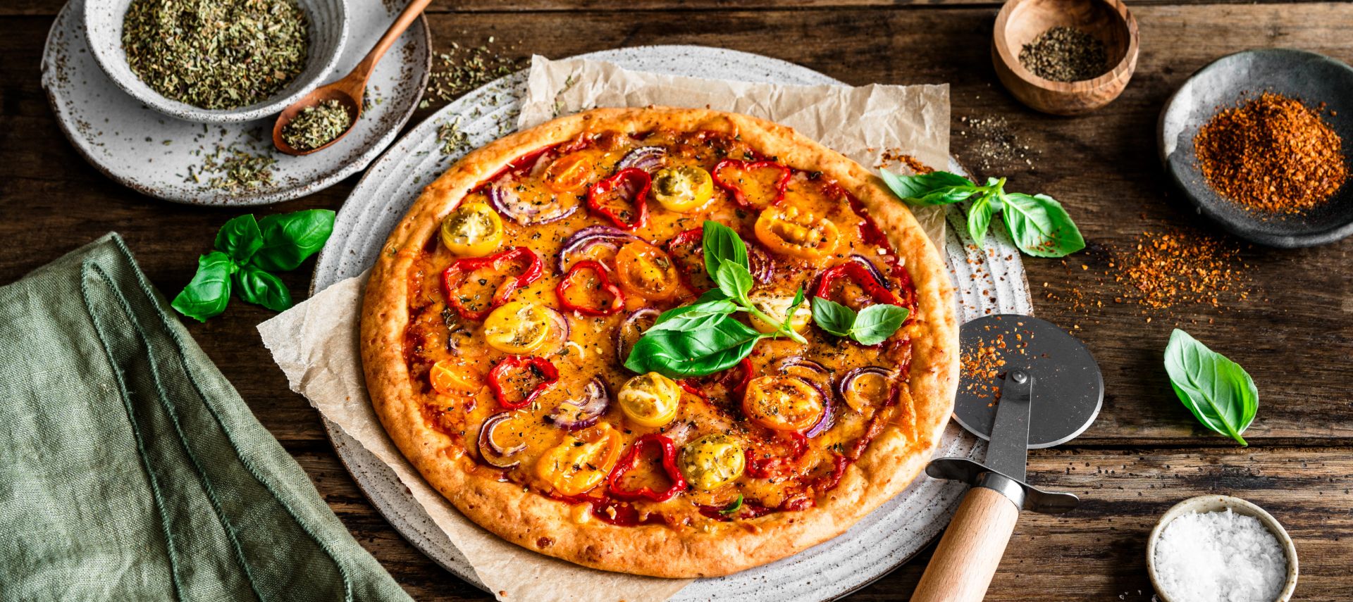 Eine hausgemachte, proteinreiche Pizza mit gelbem und rotem Tomatenbelag, Basilikumblättern und Kräutern steht auf einem rustikalen Holztisch. Daneben stehen ein Pizzaschneider, eine Serviette und kleine Schüsseln mit Gewürzen und Salz.