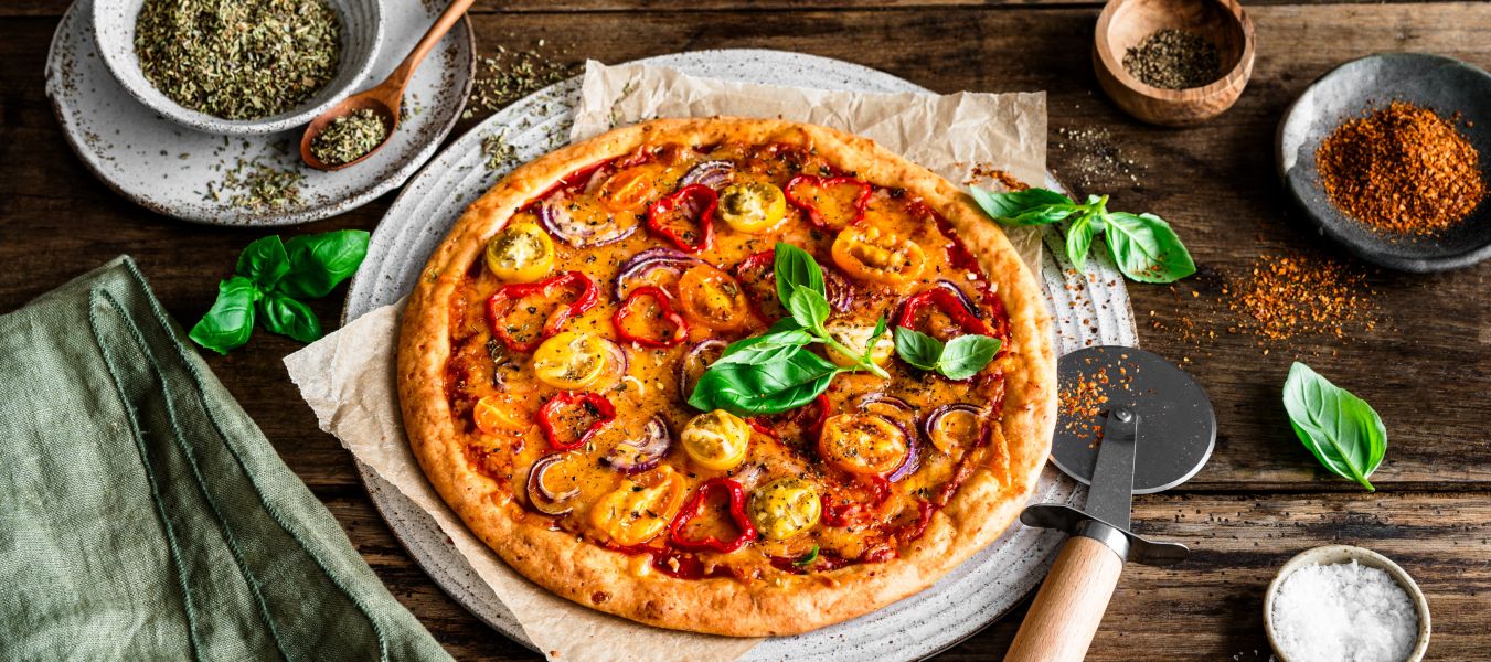 Eine hausgemachte, proteinreiche Pizza mit gelbem und rotem Tomatenbelag, Basilikumblättern und Kräutern steht auf einem rustikalen Holztisch. Daneben stehen ein Pizzaschneider, eine Serviette und kleine Schüsseln mit Gewürzen und Salz.