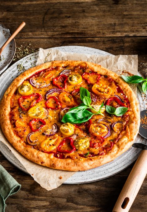 Eine hausgemachte, proteinreiche Pizza mit gelbem und rotem Tomatenbelag, Basilikumblättern und Kräutern steht auf einem rustikalen Holztisch. Daneben stehen ein Pizzaschneider, eine Serviette und kleine Schüsseln mit Gewürzen und Salz.