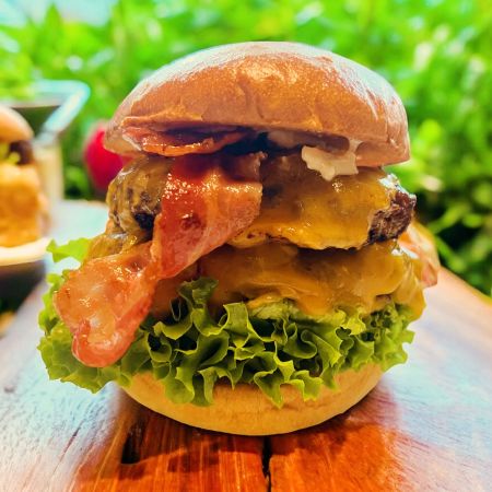 Eine Nahaufnahme eines Lippe-Cheeseburgers mit knusprigem Speck, geschmolzenem Cheddar-Käse, frischem grünem Blattsalat und einem glänzenden Brioche-Brötchen, auf einer Holzoberfläche mit unscharfem grünem Hintergrund – perfekt für jedes BBQ.