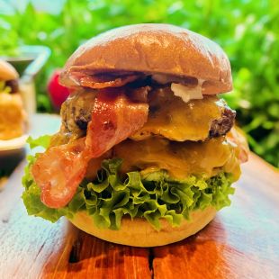 Eine Nahaufnahme eines Lippe-Cheeseburgers mit knusprigem Speck, geschmolzenem Cheddar-Käse, frischem grünem Blattsalat und einem glänzenden Brioche-Brötchen, auf einer Holzoberfläche mit unscharfem grünem Hintergrund – perfekt für jedes BBQ.