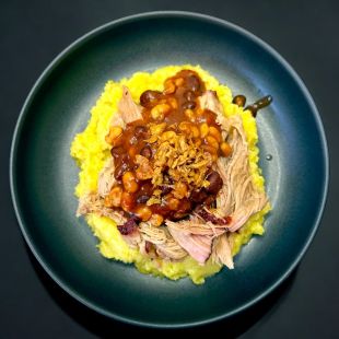 Eine Teller voll mit Kartoffelpüree garniert mit Schweinefleischstreifen, gebackenen Bohnen und knusprig gebratenen Zwiebeln, wird kunstvoll auf einem dunklen Teller angerichtet – ein unwiderstehliches Spitzengericht vor einem eindrucksvollen schwarzen Hintergrund.