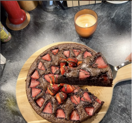 Ein Schokoladenkuchen mit Erdbeerscheiben liegt auf einem Holzbrett. Eine Hand hält einen Tortenheber und hebt ein Stück hoch, was die Freude am Kochen unterstreicht. Im Hintergrund sind eine brennende Kerze und verschiedene Küchenutensilien zu sehen.