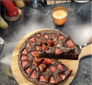 Ein Schokoladenkuchen mit Erdbeerscheiben liegt auf einem Holzbrett. Eine Hand hält einen Tortenheber und hebt ein Stück hoch, was die Freude am Kochen unterstreicht. Im Hintergrund sind eine brennende Kerze und verschiedene Küchenutensilien zu sehen.