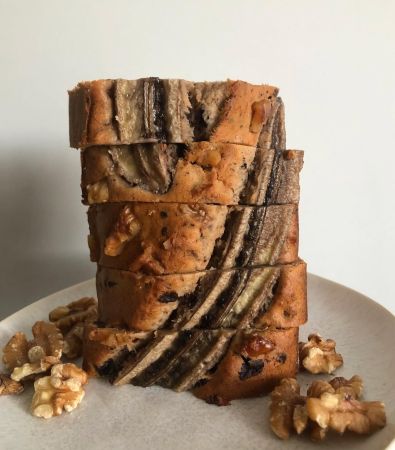 Ein Stapel dicker, köstlicher Bananenbrotscheiben mit sichtbaren Bananenscheiben und Walnüssen obenauf, angerichtet auf einem weißen Teller mit verstreuten Walnussstücken.