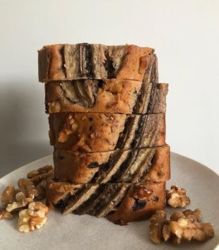 Ein Stapel dicker, köstlicher Bananenbrotscheiben mit sichtbaren Bananenscheiben und Walnüssen obenauf, angerichtet auf einem weißen Teller mit verstreuten Walnussstücken.