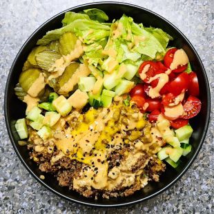 Eine Schüssel gefüllt mit Hackfleisch, garniert mit Käse und Soße, geschnittenen Essiggurken, geraspeltem Salat, gewürfelten Gurken, Kirschtomaten und noch mehr Soße ist in einzelnen Abschnitten auf einer gesprenkelten Arbeitsplatte angerichtet.