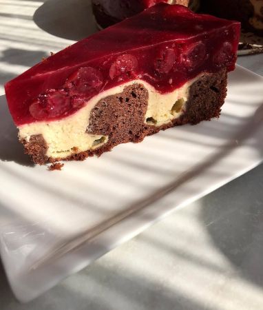 Ein Stück Schichtkuchen auf einem weißen Teller mit Schokoladenboden, cremiger Füllung mit Schokoladenstückchen und leuchtend rotem Gelee-Topping mit sichtbaren Kirschen – dieser köstliche Leckerbissen leuchtet im natürlichen Sonnenlicht.