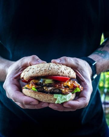 Eine Person in einem schwarzen Hemd hält einen großen, hausgemachten Joderkoch-Burger mit Salat, Tomaten, Gurken, Käse und einem Rindfleisch-Patty in einem Vollkornbrötchen und hält ihn mit beiden Händen nahe ihrer Brust.