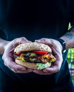 Eine Person in einem schwarzen Hemd hält einen großen, hausgemachten Joderkoch-Burger mit Salat, Tomaten, Gurken, Käse und einem Rindfleisch-Patty in einem Vollkornbrötchen und hält ihn mit beiden Händen nahe ihrer Brust.