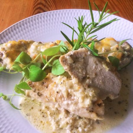 Ein weißer Teller mit cremig gebackener Hähnchenbrust, belegt mit Kräutern und grünen Microgreens, garniert mit frischem Rosmarin und serviert mit einer Käsesauce – perfekt für eine gemütliche Mahlzeit in der Kastnerischen Kantine.