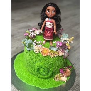 Eine Torte von bakedbyselina, dekoriert mit grünem, moosartigem Zuckerguss, bunten Blumen und Blättern, wird von einer Puppe in einem rot-weißen Kleid gekrönt, die in der Mitte steht. Die bezaubernde Torte ruht auf einer Holzoberfläche.