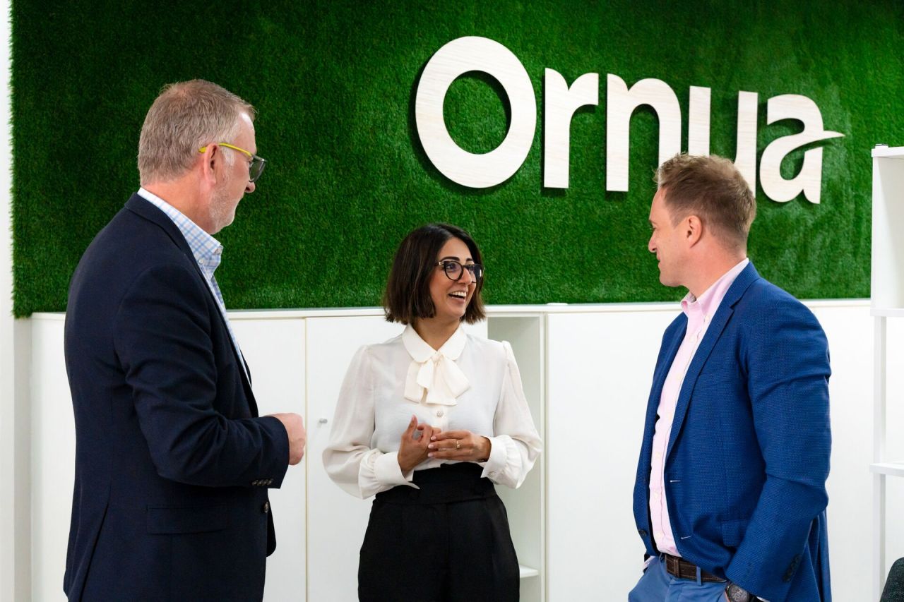 Drei Personen in Geschäftskleidung lächeln und unterhalten sich vor einer grünen Wand, auf der das Wort "Ornua" steht. Die Umgebung scheint ein modernes Büro oder ein Firmengebäude zu sein.