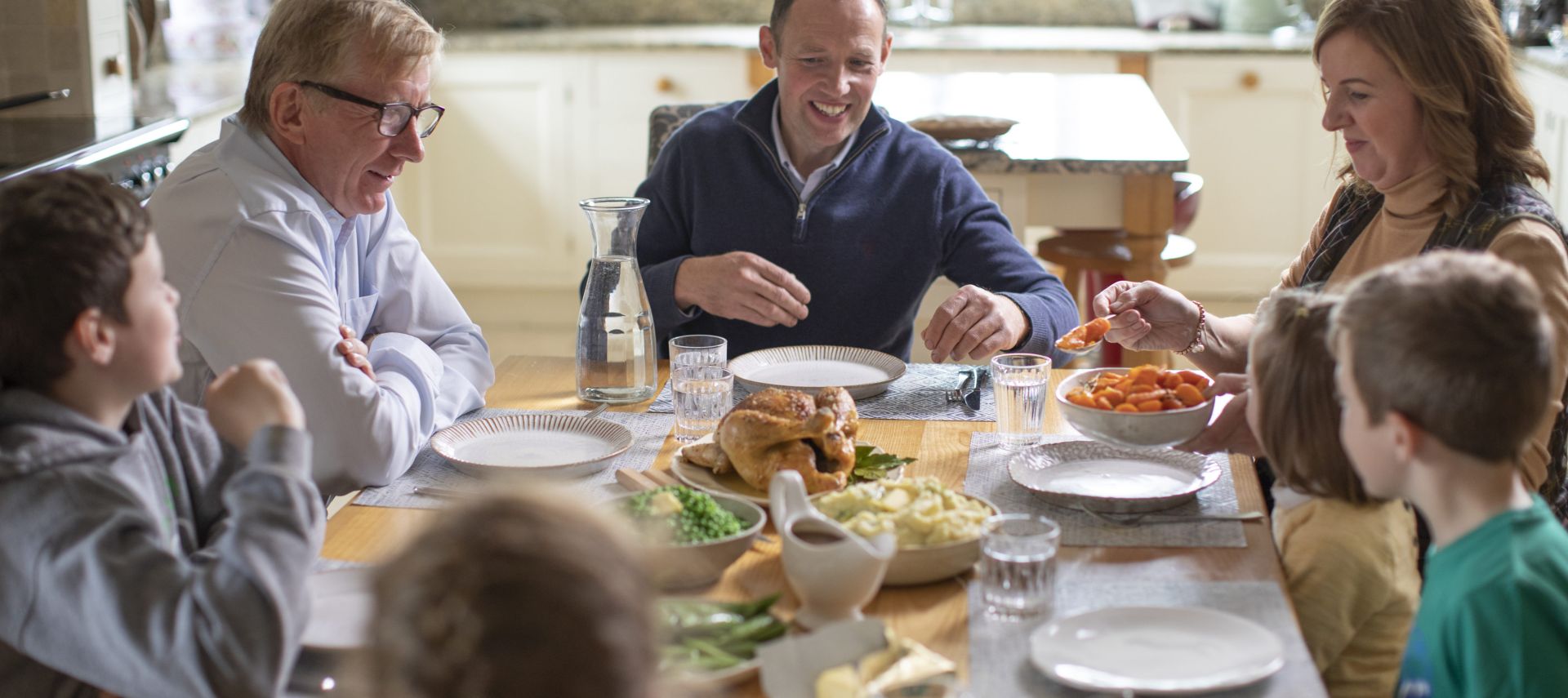 Eine Familie sitzt um einen hölzernen Esstisch, lächelt und unterhält sich, während sie das Essen serviert. Der Tisch ist mit Tellern, Gläsern, einem Brathähnchen, Gemüse und Beilagen in einer gemütlichen, sonnendurchfluteten Küche gedeckt.