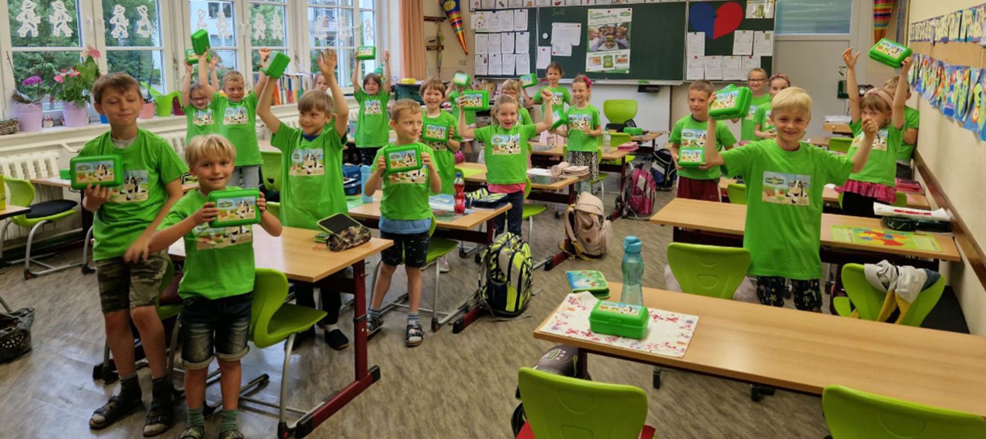 Eine Gruppe lächelnder Kinder in grünen T-Shirts steht in einem farbenfrohen Klassenzimmer und hält jeweils eine grüne Butterbrotbox hoch. Um sie herum sind Tische, Stühle, Rucksäcke und Klassenzimmerdekorationen zu sehen.