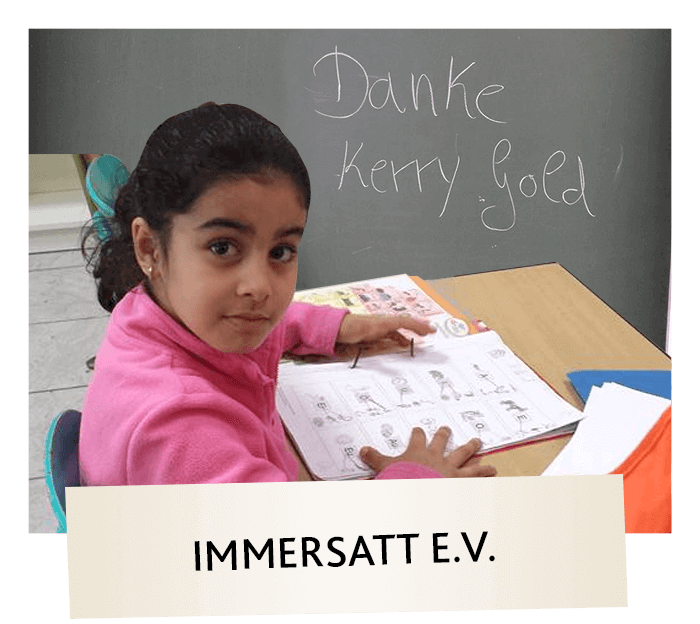 Ein junges Mädchen mit dunklen Haaren und einem rosa Pullover sitzt an einem Schreibtisch und schaut in die Kamera, während sie an einem Arbeitsbuch arbeitet. Hinter ihr ist "Danke Kerrygold" auf eine Kreidetafel geschrieben. Darunter steht der Text IMMERSATT E.V.