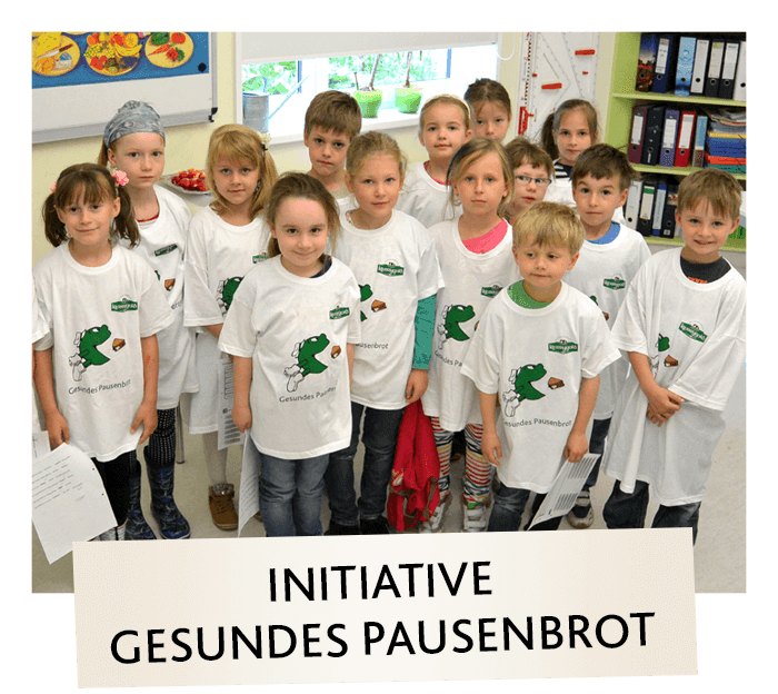 Eine Gruppe von Kindern, die weiße T-Shirts mit grünen Cartoon-Figuren tragen, steht in einem Haus zusammen und lächelt in die Kamera. Auf einem Schild davor steht INITIATIVE GESUNDES PAUSENBROT.