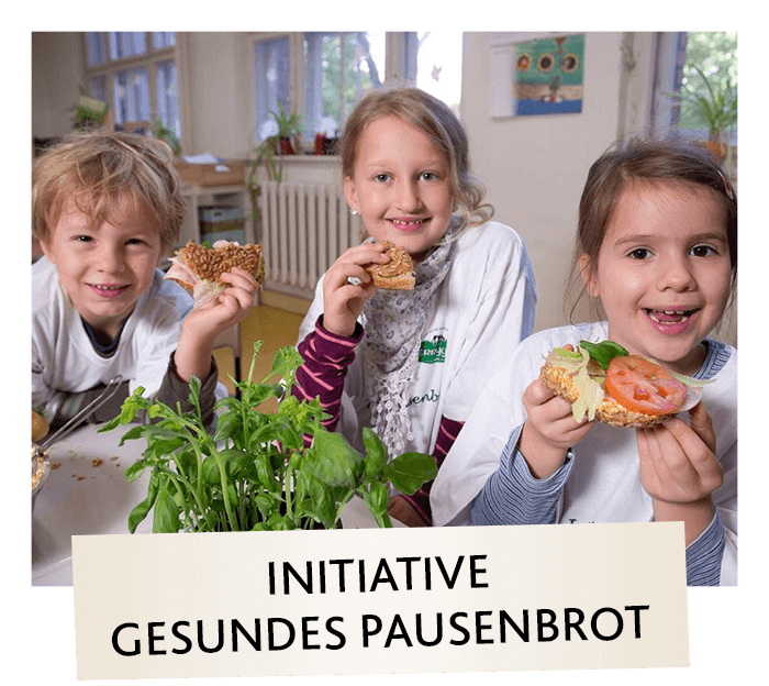Drei lächelnde Kinder sitzen an einem Tisch in einem Haus und halten gesunde Sandwiches in der Hand. Auf dem Tisch vor ihnen liegt frisches Grünzeug. Darunter steht ein Schild mit der Aufschrift INiTiATIVE GESUNDES PAUSENBROT (Initiative für einen gesunden Snack).