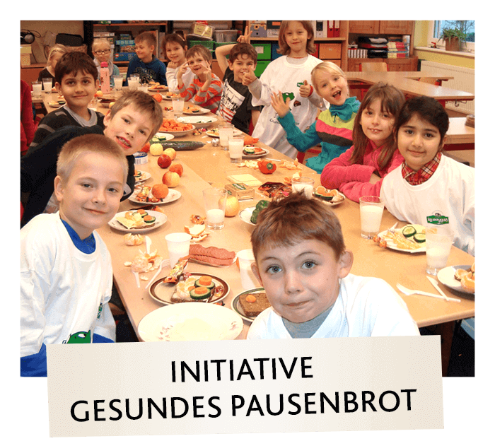 Eine Gruppe lächelnder Kinder sitzt um einen Tisch mit Tellern mit gesundem Essen, darunter Gemüse und Obst. Sie tragen weiße T-Shirts und befinden sich in einem hellen Klassenzimmer. Auf einem Schild steht "Initiative Gesundes Pausenbrot".