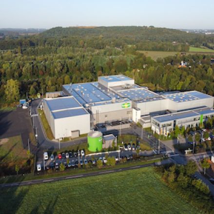 Ein großes Industriegebäude mit einem Parkplatz, umgeben von Gras, Bäumen und bewaldeten Hügeln im Hintergrund, zeigt aus der Luftperspektive das Engagement für Nachhaltigkeit.