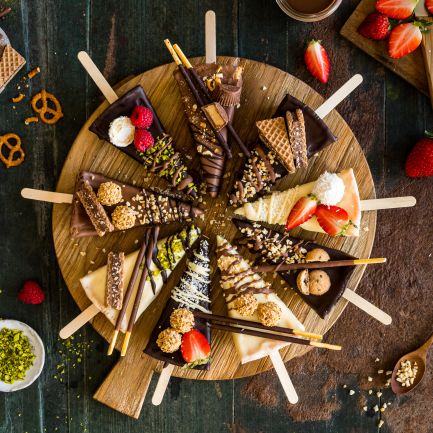 Ein runder Holzteller mit verschiedenen Dessertstücken, kreisförmig angeordnet. Jede Scheibe ist individuell dekoriert, unter anderem mit Erdbeeren, Himbeeren, Nüssen, Schokostäbchen und Waffeln. Rund um den Teller liegen weitere Zutaten und Utensilien: Erdbeeren, Himbeeren, Brezeln, gehackte Nüsse in Schälchen, ein Löffel mit Körnern oder Samen sowie ein kleines Gefäß mit Sahne oder Sauce.