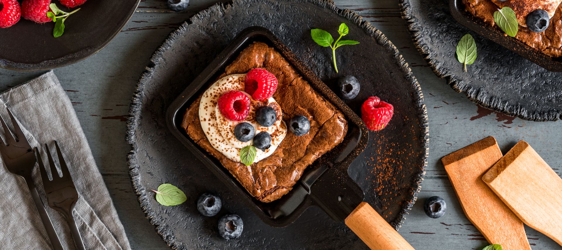 Ein quadratisches Schokoladendessert mit Schlagsahne, Himbeeren, Heidelbeeren und Minze liegt in einer kleinen Pfanne auf einem dunklen Teller, umgeben von Beeren und Utensilien auf einem rustikalen Holztisch.