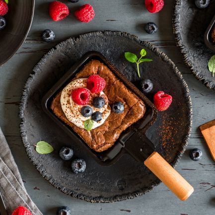 Ein quadratisches Schokoladendessert mit Schlagsahne, Himbeeren, Heidelbeeren und Minze liegt in einer kleinen Pfanne auf einem dunklen Teller, umgeben von Beeren und Utensilien auf einem rustikalen Holztisch.