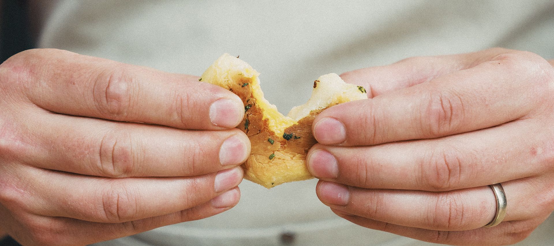 Eine Nahaufnahme von zwei Händen, die ein mit Kräutern bestreutes Stück Brot aufbrechen, das an Käsebomben erinnert. Die Person trägt einen silbernen Ring und eine helle Schürze.