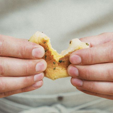 Eine Nahaufnahme von zwei Händen, die ein mit Kräutern bestreutes Stück Brot aufbrechen, das an Käsebomben erinnert. Die Person trägt einen silbernen Ring und eine helle Schürze.