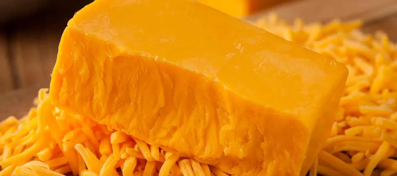Ein Block leuchtend orangefarbener Cheddar-Käse ruht auf einem Haufen zerkleinerter Cheddar-Käse auf einer hölzernen Oberfläche, mit einem anderen Käseblock verschwommen im Hintergrund.