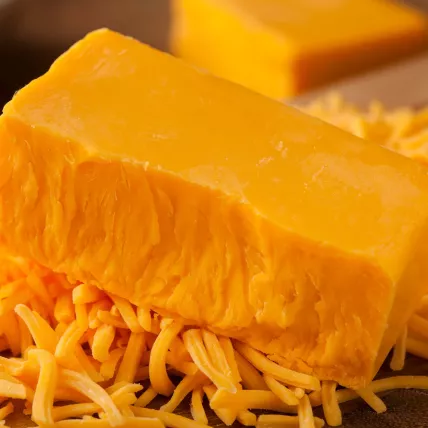 Ein Block leuchtend orangefarbener Cheddar-Käse ruht auf einem Haufen zerkleinerter Cheddar-Käse auf einer hölzernen Oberfläche, mit einem anderen Käseblock verschwommen im Hintergrund.