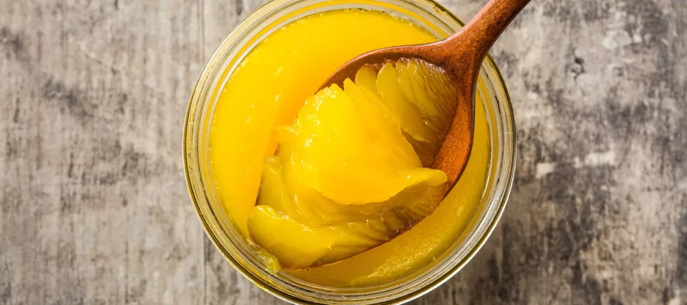 Ein Holzlöffel, der leuchtend gelbes Ghee aus einem Glasgefäß schöpft, das auf einer rustikalen Holzfläche steht - eine wesentliche Zutat in der ayurvedischen Küche.
