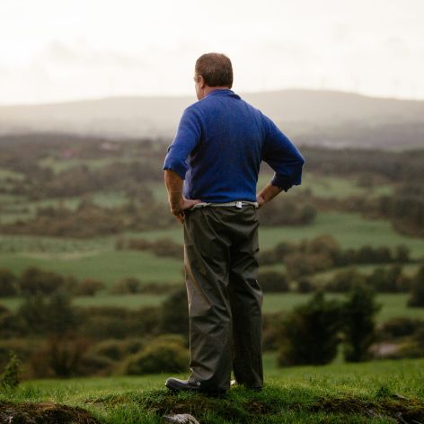 Ein Mann steht auf einem grasbewachsenen Hügel und blickt auf eine malerische Landschaft mit grünen Feldern und fernen Hügeln unter einem bewölkten Himmel.
