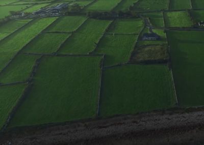 Luftaufnahme von grünen Feldern, die durch Steinzäune unterteilt sind, mit verstreuten Häusern und landwirtschaftlichen Gebäuden, die unseren Anspruch an hochwertige ländliche Landschaften widerspiegeln, umgeben von üppiger Natur.