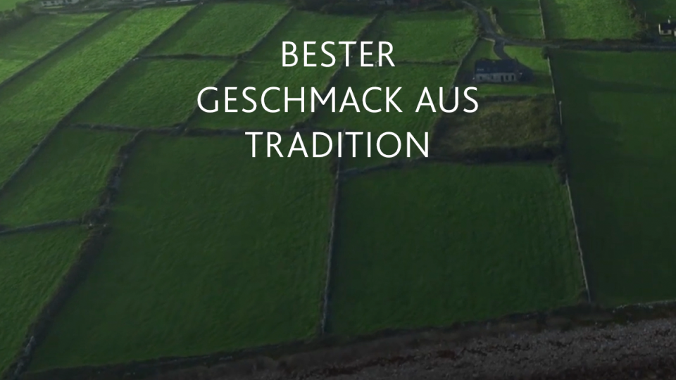 Luftaufnahme von üppig grünem Ackerland, das durch Steinmauern unterteilt ist, mit weißem Text auf Deutsch: Bester Geschmack aus Tradition.