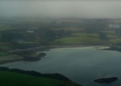 Eine neblige Landschaft mit einem ruhigen Gewässer, umgeben von grünen Hügeln und Feldern, spiegelt unseren Anspruch auf heitere Schönheit wider. Der Hintergrund ist in Nebel gehüllt, der die entfernten Gebäude verdeckt und den Horizont mit dem wolkenverhangenen Himmel verschmelzen lässt.