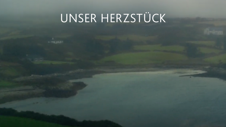 Eine neblige Küstenlandschaft mit grünen Feldern, felsiger Küste und ruhigem Wasser unter einem bewölkten Himmel. Der deutsche Text UNSER HERZSTÜCK erscheint in der Mitte des Bildes.