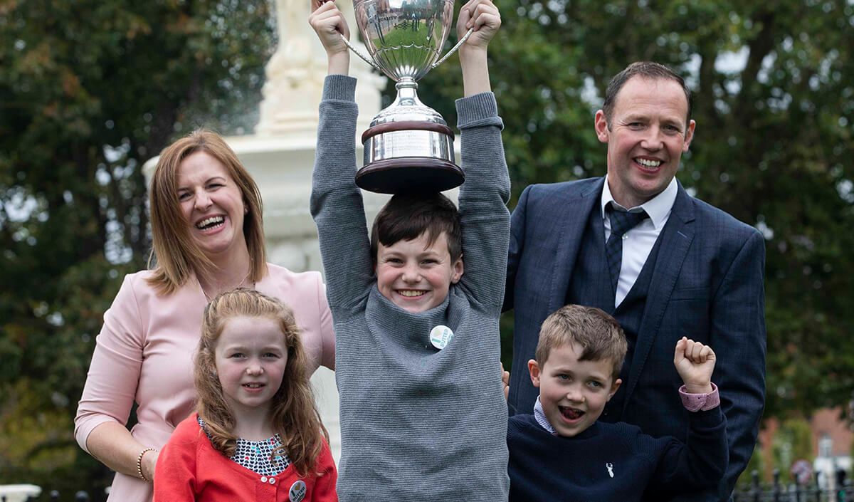 Ein lächelnder Junge hält eine Trophäe hoch, während sich drei Kinder und zwei Erwachsene im Freien um ihn versammeln. Die Gruppe sieht glücklich und feierlich aus, im Hintergrund sind Bäume und ein weißer Brunnen zu sehen.