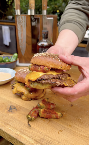 Eine Person hält einen Bergnbeef-Cheeseburger mit Cheddar-Käse, mit Speck umwickelten Jalapeños und einem Sesambrötchen über einem hölzernen Schneidebrett mit weiteren Burgern und gefüllten Jalapeños.