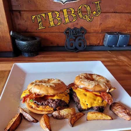 Zwei Cheeseburger mit Speck auf einem quadratischen weißen Teller, serviert mit Kartoffelspalten. Das rustikale Holzdekor mit einem Route-66-Schild und dem Schriftzug TBBQJ an der Wand schafft eine authentische Atmosphäre.