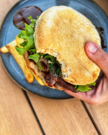 Eine Hand hält einen angebissenen Burger mit Salat und Fleisch, über einem Teller mit Pommes frites und dunkler Dip-Sauce auf einem Holztisch.