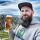 Ein bärtiger Mann mit Mütze und Kapuzenpulli hält ein Glas Bier in der Hand und lächelt, während er draußen steht. Im Hintergrund sind Berge und ein wolkenverhangener blauer Himmel zu sehen.