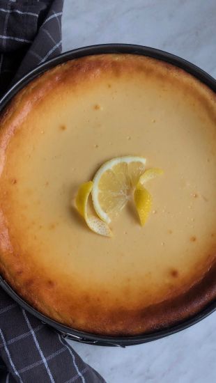 Ein xbambilicious gebackener Käsekuchen mit goldbraunem Rand, der mit zwei Zitronenscheiben belegt ist, steht auf einer Marmorplatte neben einem dunkel karierten Tuch.