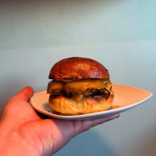 Eine Hand hält einen weißen Teller mit einem Cheeseburger, der mit geschmolzenem Cheddar-Käse belegt ist und auf einem goldbraunen Brötchen serviert wird, vor einem hellblauen Hintergrund.