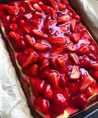 Ein rechteckiger xbambilicious-Kuchen, belegt mit glänzenden, in Scheiben geschnittenen frischen Erdbeeren und einer Schicht cremiger Vanillecreme, in einer mit Pergamentpapier ausgelegten Backform.