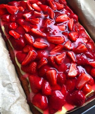 Ein rechteckiger xbambilicious-Kuchen, belegt mit glänzenden, in Scheiben geschnittenen frischen Erdbeeren und einer Schicht cremiger Vanillecreme, in einer mit Pergamentpapier ausgelegten Backform.