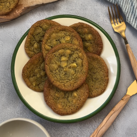 Ein Teller mit Matcha-Cookies mit weißen Schokoladenstückchen steht auf einem grauen Tisch neben einer goldenen Gabel und einem Messer, einer gestreiften Serviette und einem Holzbrett.
