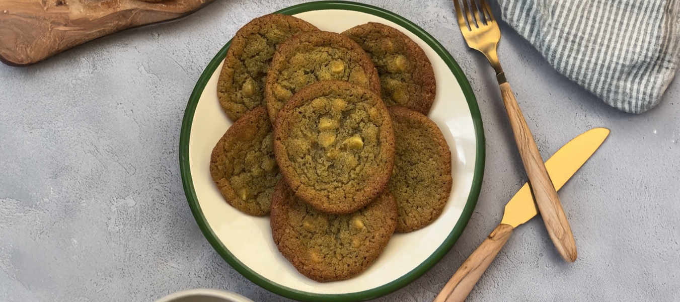 Ein Teller mit Matcha-Cookies mit weißen Schokoladenstückchen steht auf einem grauen Tisch neben einer goldenen Gabel und einem Messer, einer gestreiften Serviette und einem Holzbrett.
