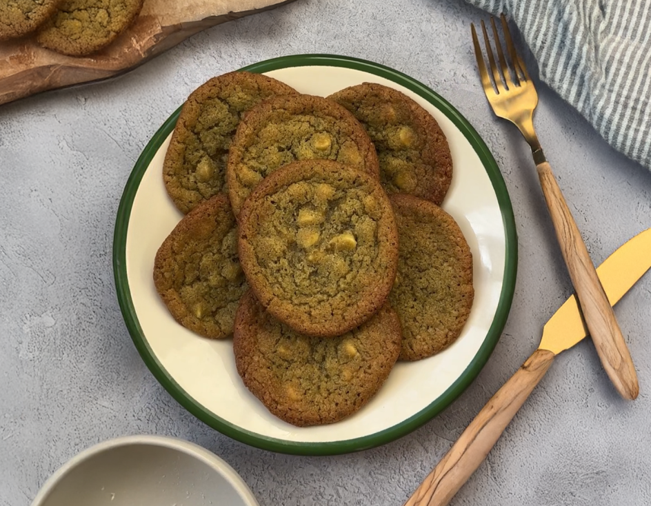 Ein Teller mit Matcha-Cookies mit weißen Schokoladenstückchen steht auf einem grauen Tisch neben einer goldenen Gabel und einem Messer, einer gestreiften Serviette und einem Holzbrett.